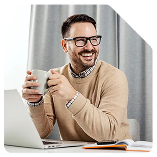 A happy man in smart attire sat in front of a laptop holds a coffee cup